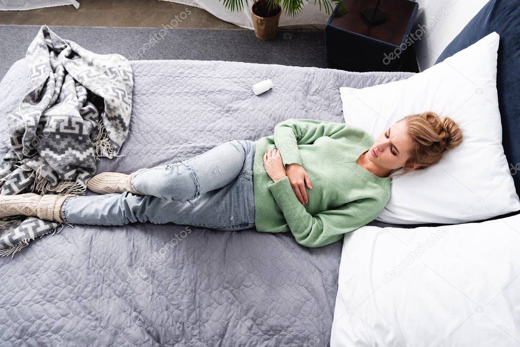 top view of woman having stomachache and lying on bed with bottle of pills