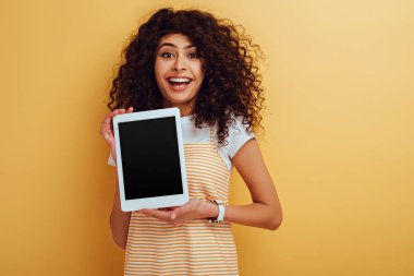 Sarı arka planda boş ekranlı dijital tablet gösteren neşeli iki ırklı kız.