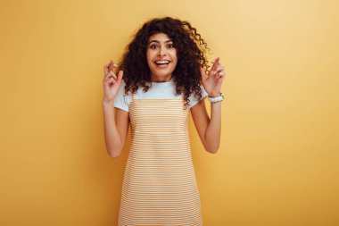 excited bi-racial girl holding crossed fingers while smiling at camera on yellow background clipart