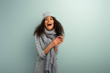 excited bi-racial girl in warm hat and scarf laughing on grey background clipart