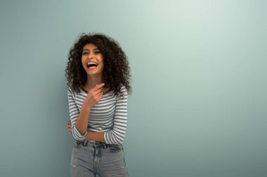 excited bi-racial girl laughing while pointing with finger on grey background clipart