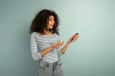 displeased mixed race girl showing question gesture while looking at smartphone on grey background clipart