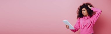 Pembe arka plandaki dijital tablete bakarken saçına dokunan hayal kırıklığına uğramış çift ırklı kızın panoramik görüntüsü.