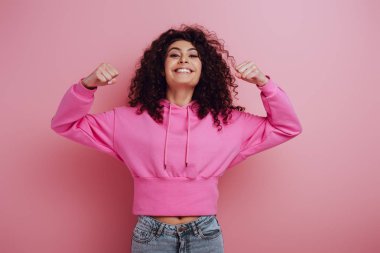 excited bi-racial girl showing winner gesture while smiling at camera on pink background clipart