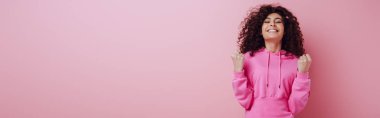 panoramic shot of excited bi-racial girl showing winner gesture while standing with closed eyes on pink background clipart