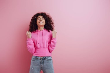 excited bi-racial girl showing winner gesture while standing with closed eyes on pink background clipart