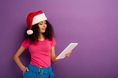 Gülümseyen çift ırklı Noel Baba şapkalı kız mor arka planda dijital tablet kullanıyor.