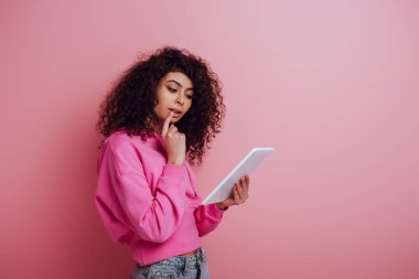 Pembe arka planda dijital tablet kullanırken dudağa dokunan düşünceli iki ırklı kız.