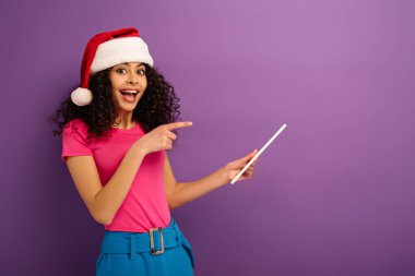 Noel Baba şapkalı neşeli melez kız mor arka planda dijital tableti işaret ediyor.