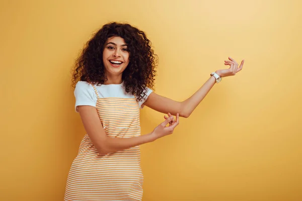 黄色い背景のカメラで微笑みながら手を指差す陽気なバイレーシャルの女の子 — ストック写真