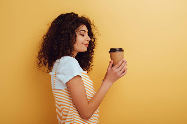 вид сбоку улыбающейся девушки смешанной расы, держащей кофе на желтом фоне

