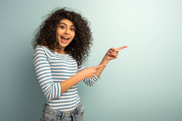 excited mixed race girl looking at camera while pointing with fingers on grey background