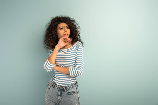 вдумчивая двурасовая девушка, держащаяся за руки возле лица, отводя взгляд на сером фоне
