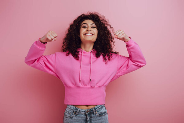 excited bi-racial girl showing winner gesture while smiling at camera on pink background