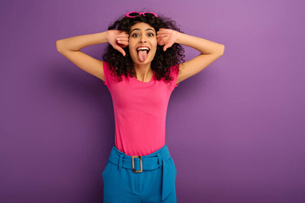 cheerful bi-racial girl sticking out tongue while looking at camera on purple background