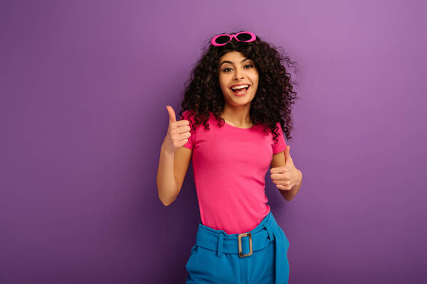 cheerful bi-racial girl showing thumbs up while smiling at camera on purple background