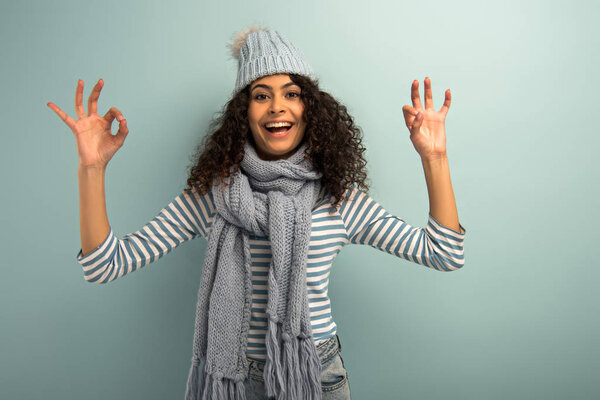 happy bi-racial girl in warm hat and scarf showing okay signs while looking at camera on grey background