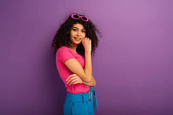 attractive bi-racial girl smiling at camera while holding hand near face on purple background