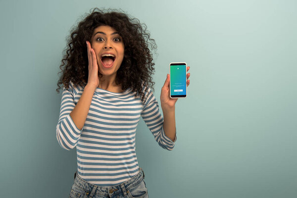 1KYIV, UKRAINE - NOVEMBER 26, 2019: excited bi-racial girl looking at camera while showing smartphone with Twitter app on grey background