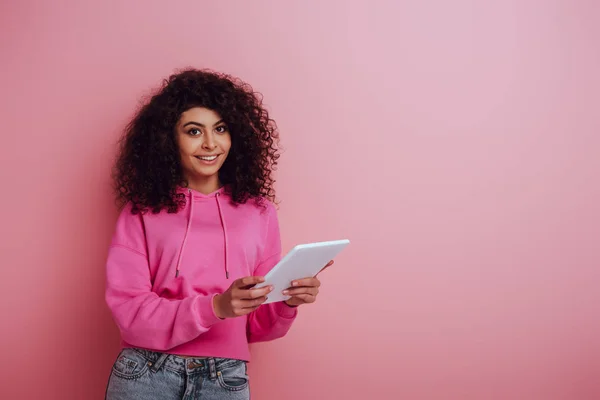 快乐的混血女孩一边在镜头前微笑 一边用粉色背景的数码平板电脑 — 图库照片