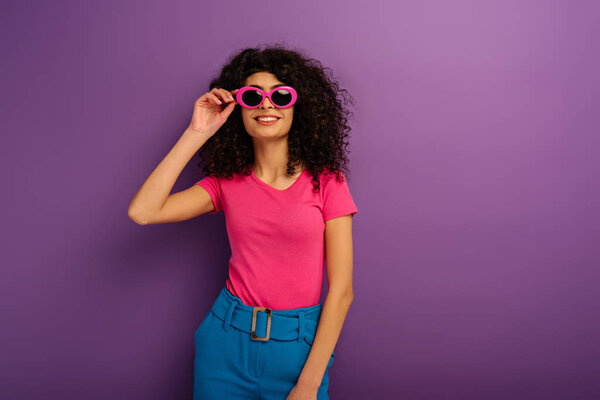 cheerful bi-racial girl touching sunglasses while looking at camera on purple background