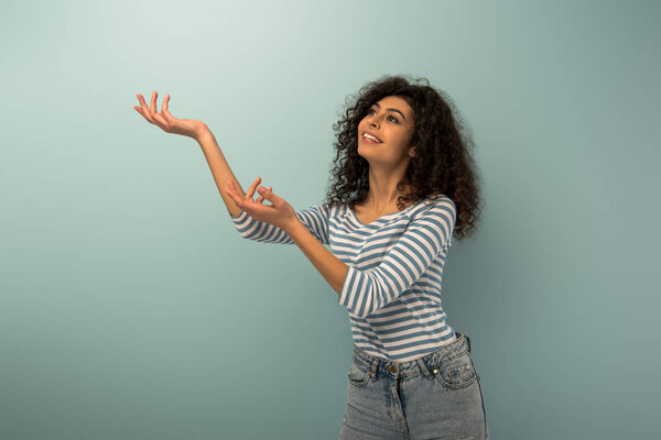 улыбающаяся двурасовая девушка смотрит вверх, стоя с протянутыми руками на сером фоне
