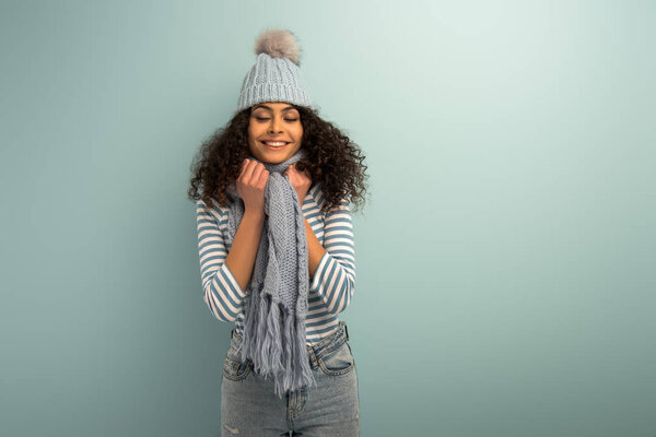 pretty bi-racial girl in warm hat and scarf smiling with closed eyes on grey background