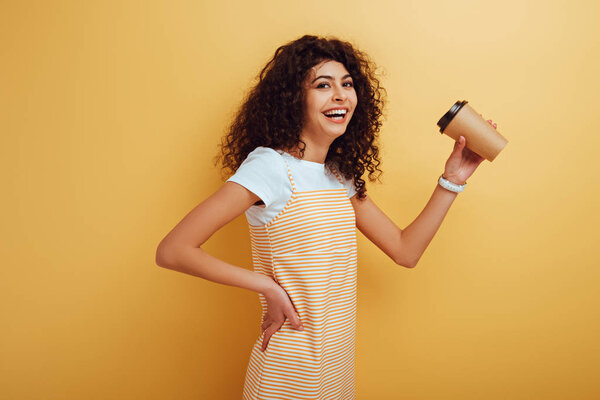 веселая двурасовая девушка улыбается в камеру, держа кофе на желтом фоне
