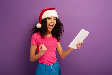 Mor arka planda dijital tablet tutarken kazanan jesti gösteren Noel Baba şapkalı heyecanlı melez kız