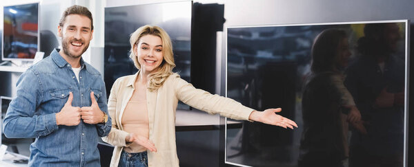 panoramic shot of boyfriend showing likes and smiling girlfriend pointing with hand at new tv in home appliance store 