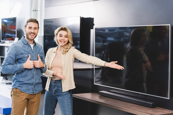 boyfriend showing likes and smiling girlfriend pointing with hand at new tv in home appliance store 