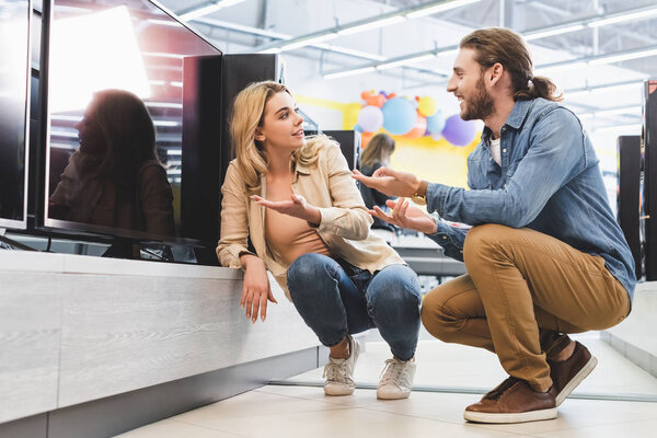 Улыбающиеся бойфренд и девушка указывая руками на новый телевизор и говорить в магазине бытовой техники
 