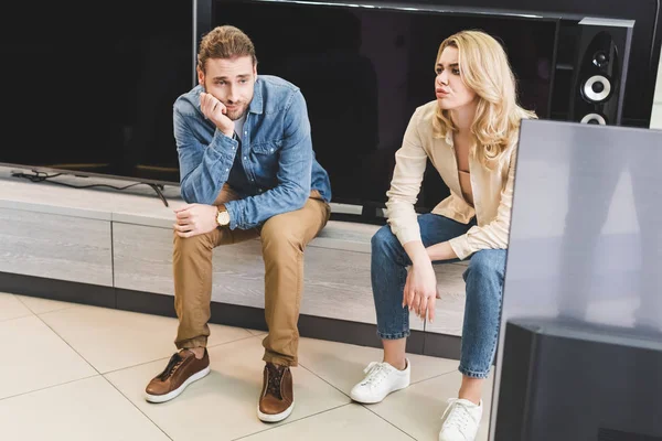家電店の新しいテレビの近くに座っている悲しいボーイフレンドとガールフレンド — ストック写真