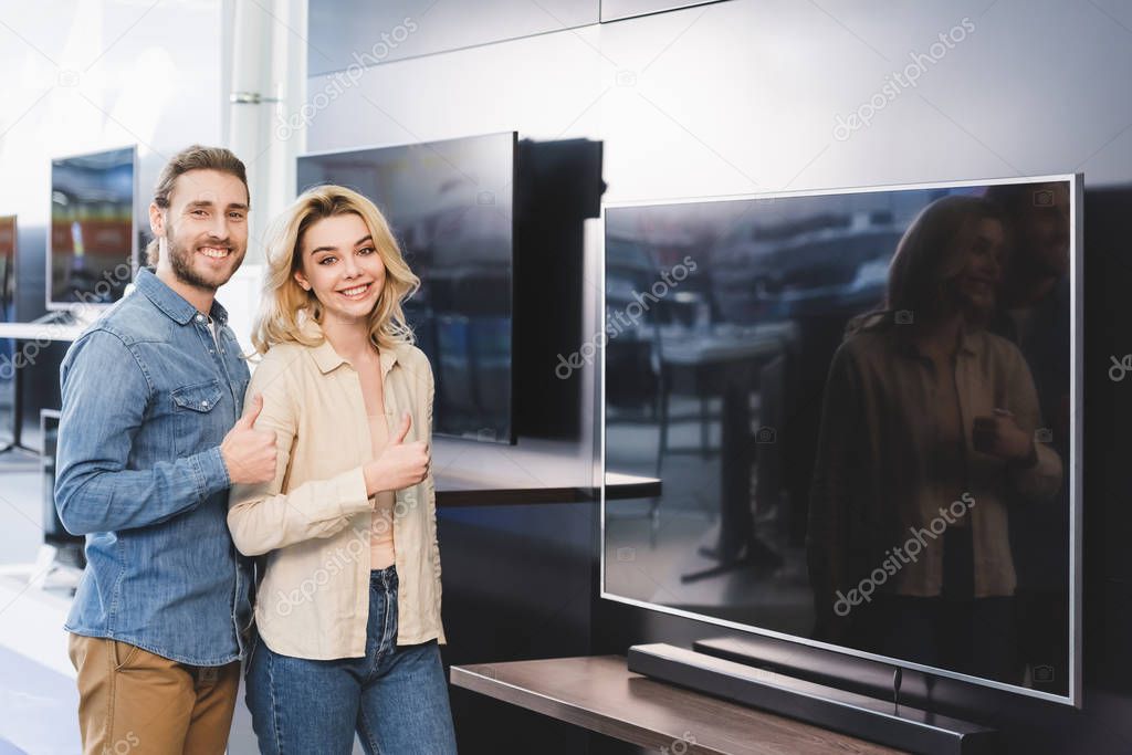 smiling boyfriend and girlfriend showing thumbs up in home appliance store 