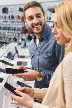 Gülümseyen erkek arkadaşın ev aletleri mağazasında yeni akıllı telefonlu kız arkadaşına bakması. 