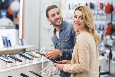 smiling boyfriend and girlfriend holding new smartphones in home appliance store  clipart