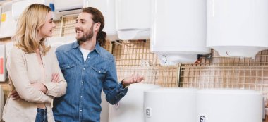 panoramic shot of smiling boyfriend and girlfriend talking near boilers in home appliance store  clipart