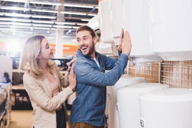 smiling boyfriend touching boiler and looking at girlfriend in home appliance store  clipart