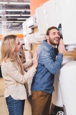 smiling boyfriend touching boiler and girlfriend looking at him in home appliance store  clipart