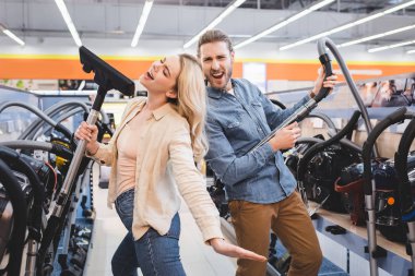 boyfriend playing with vacuum cleaner and girlfriend singing in home appliance store  clipart