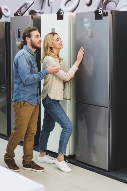 boyfriend pointing with hand and smiling girlfriend touching fridge in home appliance store  clipart