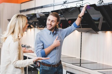 smiling consultant pointing with hand at cooker hood and talking with shocked woman in home appliance store  clipart
