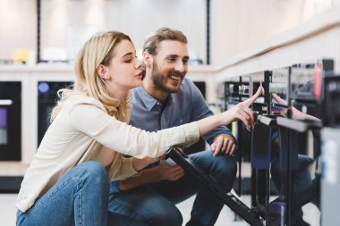 woman using oven and smiling consultant looking at it in home appliance store  clipart