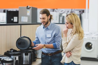 smiling consultant pointing with hand at slow cooker and pensive woman looking at it in home appliance store  clipart