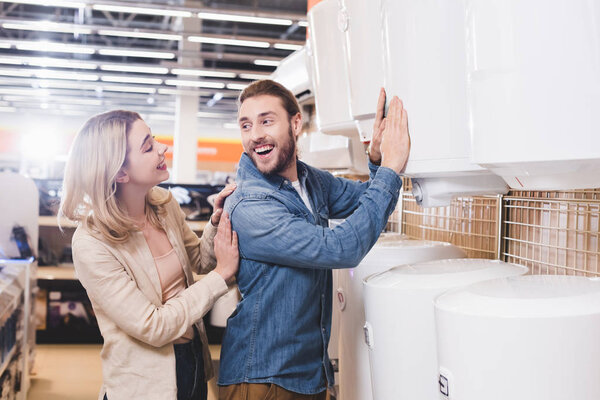smiling boyfriend touching boiler and looking at girlfriend in home appliance store 