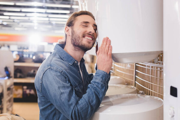 smiling man with closed eyes touching boiler in home appliance store 