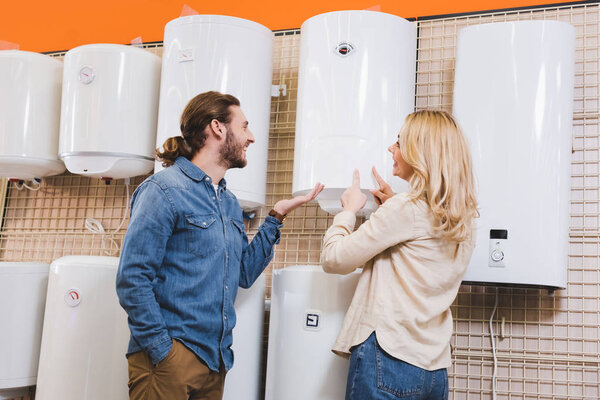 smiling boyfriend pointing with hand and girlfriend pointing with fingers at boiler in home appliance store 