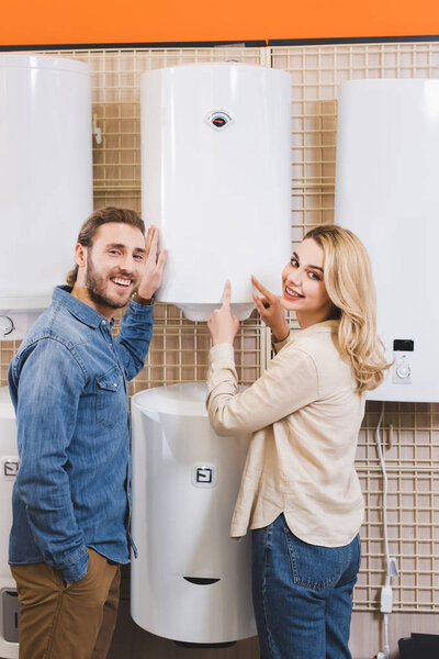 smiling boyfriend and girlfriend pointing with fingers at boiler in home appliance store 