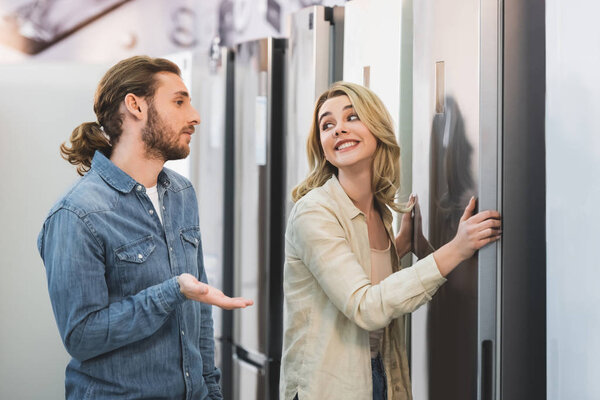boyfriend talking and smiling girlfriend touching fridge and looking at him in home appliance store 