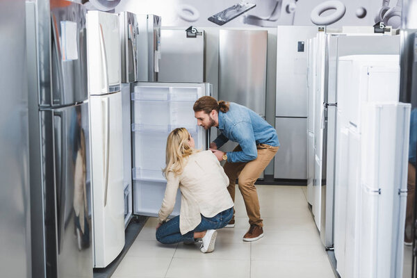 boyfriend and girlfriend talking near opened fridge in home appliance store 